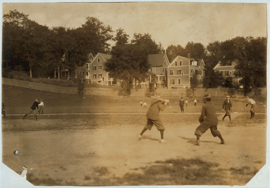 Young people play baseball on an open playground. A building and utility poles rise beyond the rough field.